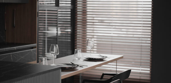 Mimika(Мимика). Просторная фотостудия. Современное помещение с деревянным столом у окна с жалюзи. На столе сервировка для двух человек: тарелки, бокалы, приборы. Два черных стула, темные деревянные элементы, светлая и серая гамма интерьера.