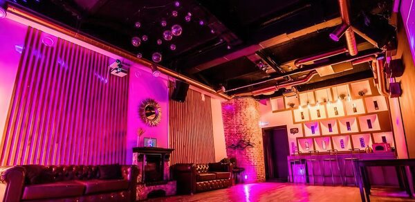 LoftForParty. Бэкстейдж лофт . Тёмное помещение с низким потолком, кожаные диваны, барная стойка, разноцветное освещение, стены отделаны деревянными панелями и кирпичом, металлические трубы на потолке, зеркальный декор, дискоболы, деревянный пол.