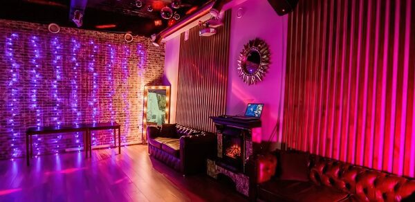 LoftForParty. Бэкстейдж лофт . Кирпичная стена с фиолетовой подсветкой, пластиковые стулья, диваны из тёмной кожи, деревянный пол, подвесной светильник, зеркало с подсветкой, декоративное зеркало на стене, мебель и стены в теплых тёмных тонах.