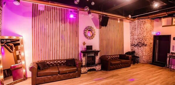 LoftForParty. Бэкстейдж лофт . Простор с деревянным полом и светлыми стенами, декорированными вертикальными панелями. Две коричневые кожаные софы, зеркало, настенное декоративное зеркало, несколько подвесных диско-шаров, сцена с DJ-аппаратурой, разноцветное освещение.
