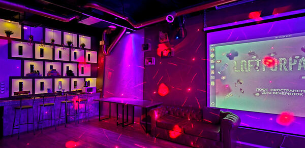 LoftForParty. Бэкстейдж лофт . Арендное пространство с низким баром и стульями, настроенной полкой с подсветкой. Рядом находится диван у стены с проекционным экраном. Освещение разноцветное, пол деревянный. Стены тёмные с декоративными элементами.