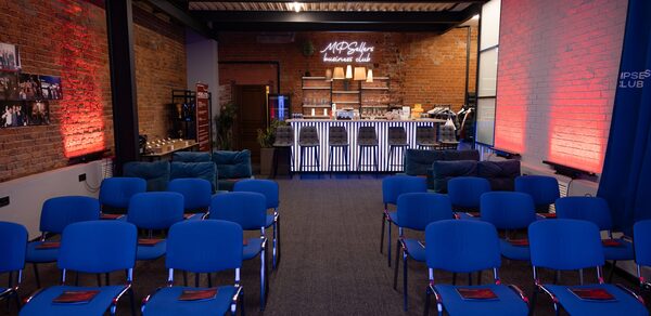 MPSellers(МПСеллерс). Просторный зал для мероприятий. Пространство с кирпичными стенами, металлической крышей и синим освещением. В центре ряды синих стульев, напротив барная стойка с неоновым знаком. Справа белая стена с красной подсветкой. Слева фотографии на стене.