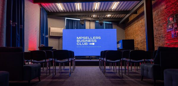 MPSellers(МПСеллерс). Просторный зал для мероприятий. Пространство в стиле лофт с кирпичными стенами и металлическим потолком, с проектором и экраном. Ряд черных кресел, синий занавес, разноцветная подсветка. Надпись на экране: "MPSELLERS BUSINESS CLUB".