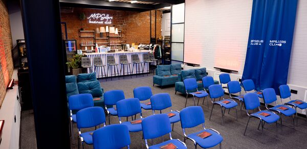 MPSellers(МПСеллерс). Просторный зал для мероприятий. Пространство зала с рядами голубых стульев и мягкой мебелью у кирпичной стены. Барная стойка с высокими стульями, задник из белого кирпича. Синий занавес с надписями. Металлические балки на потолке, ковровое покрытие. Надпись на стене: "MPG..."
