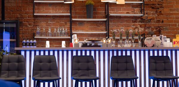 MPSellers(МПСеллерс). Просторный зал для мероприятий. Барная стойка с высокими черными стульями, кирпичная стена, полки с бутылками и бокалами, несколько ламп с абажурами. Бело-голубая подсветка стойки, на полке растение в горшке.