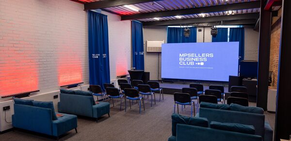 MPSellers(МПСеллерс). Просторный зал для мероприятий. Аудитория с проектором и экраном, расстановкой стульев в нескольких рядах. Несколько диванов сзади, синие и красные светодиодные светильники на кирпичных стенах, синие театральные шторы, металлический потолок, серый ковролин, кондиционер на стене.