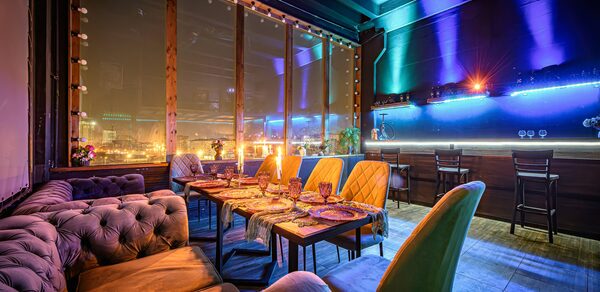 Лофт ZUM(Зум). Полузатемнённое помещение с панорамными окнами, обставленное мягкими диванами и стульями. Длинный стол покрыт дорожкой, на которой расставлены бокалы и тарелки. Яркое разноцветное неоновое освещение, стены сине-фиолетовые, пол серый.