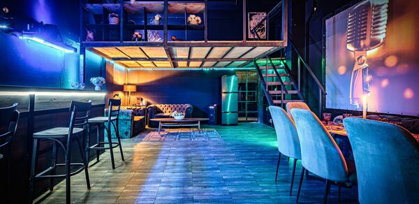 Лофт ZUM(Зум). Просторное помещение с диванами и креслами, барной стойкой и высокими стульями. На стенах неоновая подсветка и проектор с изображением микрофона. Лестница ведет на второй уровень. Напольное покрытие из плитки, освещение мягкое, разноцветное.