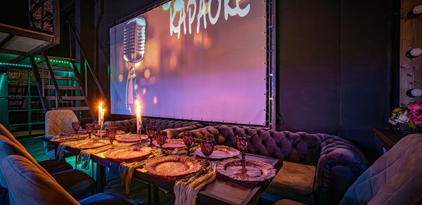 Лофт ZUM(Зум). Караоке-зал с приглушенным светом, длинный деревянный стол с сервировкой для вечеринки, бархатные диваны и стулья. На стене большой экран с изображением микрофона и надписью "Караоке". Подсвеченные лестницы и светодиодные огни.