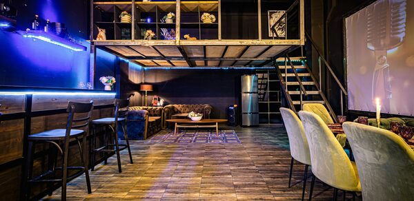 Лофт ZUM(Зум). Лофт-пространство с открытым вторым уровнем, лестницей, барной стойкой с высокими стульями, диванами, деревянным столом. Деревянный пол, мягкие стулья, декоративные огни, металлический каркас. Тёмные стены, яркое освещение, металлический холодильник.