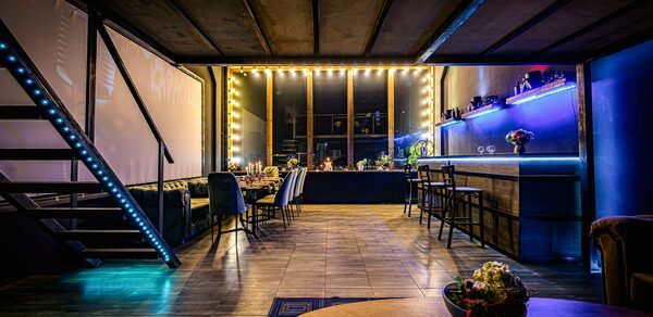 Лофт ZUM(Зум). Лофт-пространство с лестницей, вдоль стены большой стол с черными стульями, освещение от гирлянд и светодиодов. Барная стойка с высокими стульями, пол деревянный, потолок из темных панелей. Синие и золотые тона, присутствуют цветы.