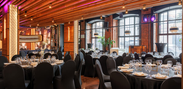 Zafferano Loft(Зафферано лофт). Банкетный зал с круглым столами, покрытыми черными скатертями и сервировкой из бокалов и тарелок. Кресла в черных чехлах. Потолок с деревянными балками и встроенным освещением. Стены из кирпича с большими арочными окнами.