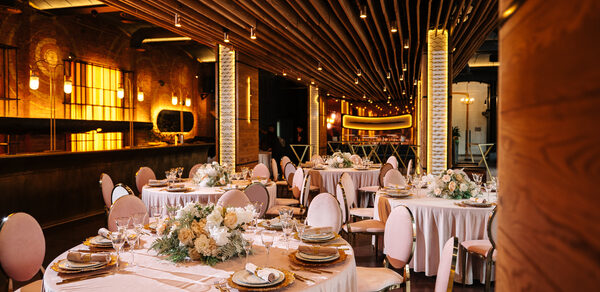 Zafferano Loft(Зафферано лофт). Банкетный зал с круглым столами, покрытыми белыми скатертями, украшенными цветочными композициями. Розовые стулья, темное деревянное потолочное покрытие с встроенными светильниками. Барная стойка с золотистой подсветкой, кирпичные стены.