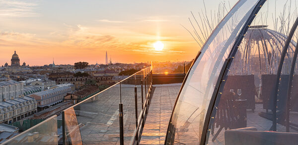 Sky Terrasse(Небо Терраса). Терраса с панорамным видом на город, ограждена стеклянными перилами. Справа расположена прозрачная конструкция с плетеной мебелью внутри, украшенной гирляндами. Закатное небо с ярким солнцем. Цвета: оранжевый, голубой, серый.