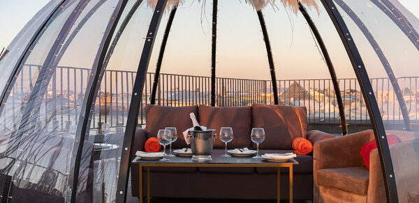 Sky Terrasse(Небо Терраса). Прозрачная геодезическая палатка на террасе. Внутри коричневая мягкая мебель: диван и кресло. Перед ними стол с белыми тарелками, бокалами и ведёрком для шампанского. Декор сверху из сухих растений. Железные перила на заднем плане.