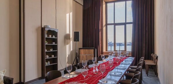 Chocolate Loft(Шоколад Лофт). Зал с видом на Финский залив. Просторное помещение с длинным столом и стульями. На столе красная дорожка, стеклянные бокалы и тарелки. Большое окно с тёмными занавесками. На стенах полки с посудой, акустическая колонка и проектор. Серые полы, светлые стены, подвесное освещение.