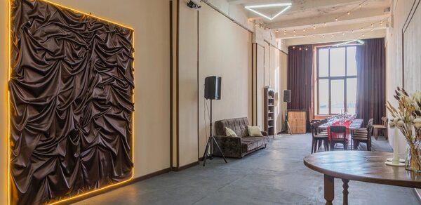 Chocolate Loft(Шоколад Лофт). Зал с видом на Финский залив. Просторное помещение с высоким потолком, белые стены, на одной из стен драпировка с подсветкой. Большое окно с темными шторами, стол и стулья, коричневый диван. Стена с колонками, деревянный круглый стол. Светодиодный светильник треугольной формы на потолке.