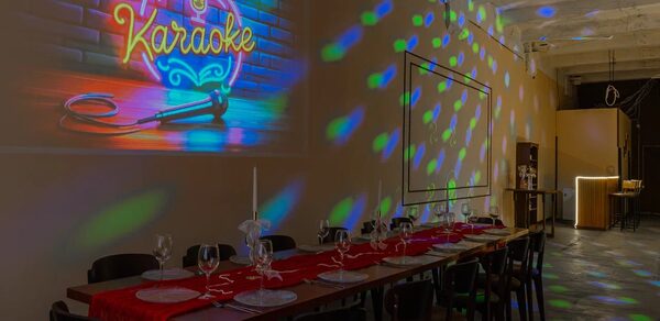 Chocolate Loft(Шоколад Лофт). Зал с видом на Финский залив. Пространство с длинным столом, покрытым красной дорожкой, сервировано бокалами и приборами. На кирпичной стене вывеска "Karaoke" с неоновыми микрофоном. Потолок высокий, с диско-освещением. Справа стойка бара, помещение в темных тонах.