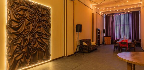 Chocolate Loft(Шоколад Лофт). Зал с видом на Финский залив. Пространство с высокими потолками, длинный стол с красными стульями, диван, деревянный круглый стол. Темные шторы, скульптурное панно с подсветкой, гирлянды на потолке. Светлые стены, темный пол, колонны. Освещение теплое, атмосферное.