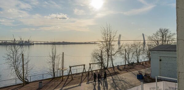 Chocolate Loft(Шоколад Лофт). Зал с видом на Финский залив. Пространство на набережной с видом на водоём и мост. Пешеходная дорожка с деревьями и уличным освещением. Светлое время суток, солнечно. Люди гуляют. Вдали видны портовые краны. Слева ограждение со знаками.