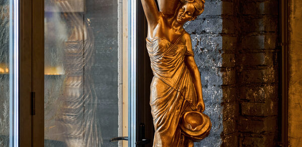 Tangiers Lounge(Танжер Лаундж) . Золотая статуя женщины в классическом стиле рядом с высоким стеклянным окном. Кирпичная стена тёмного цвета на заднем плане. Мягкое освещение подсвечивает детали скульптуры и текстуру стены.