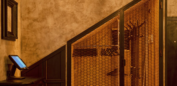 Tangiers Lounge(Танжер Лаундж) . Помещение с кирпичными стенами и деревянной отделкой. Под лестницей металлическая решетка; рядом темная деревянная мебель с экраном. Потолочный светильник с теплым освещением. Стены обшиты панелями, общей цвет — теплый, коричневый.