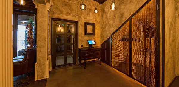 Tangiers Lounge(Танжер Лаундж) . Интерьерное пространство с приглушенным освещением, бетонными стенами и потолком. Тёмная дверь с стеклянной вставкой, небольшой стол с экраном. Деревянная решётчатая перегородка, подвесные светильники. Отделка из дерева и металла, нейтральные тона.