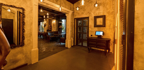 Tangiers Lounge(Танжер Лаундж) . Стильное пространство с антикварными элементами: арочная ниша, зеркала в резных рамах, плиточный пол, приглушённое тёплое освещение с подвесными лампами, деревянный стол с монитором. Стены с фактурной отделкой, некоторые декорированы пустыми рамками.