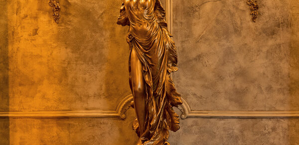 Tangiers Lounge(Танжер Лаундж) . Золотая скульптура девушки с поднятыми руками на фоне стены с декоративной штукатуркой серо-бежевого цвета, украшенной лепнинами. По бокам стены расположены две настенные лампы в классическом стиле с резными элементами. Освещение теплое.