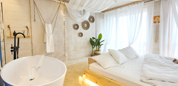 Пляжный лофт с галькой, открытой ванной, панорамными окнами. Просторная светлая зона с ванной и кроватью. Ванная белая, отдельно стоящая. Кровать с деревянным каркасом, белое постельное белье. Светло-бежевые стены с декоративными элементами. Растение в углу, на окнах белые шторы, потолочные тканевые занавеси.