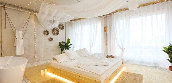 Пляжный лофт с галькой, открытой ванной, панорамными окнами. Просторное светлое помещение с кроватью на деревянном подиуме, подсветка под кроватью, белые шторы, подвешенный прозрачный балдахин. Ванна отдельно, круглая, белого цвета. Декоративные элементы на стене. Паркетный пол, две плетёные коврики.