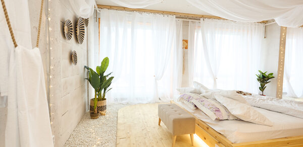 Пляжный лофт с галькой, открытой ванной, панорамными окнами. Светлое помещение с деревянным полом и гравийным покрытием. Большая кровать с декоративными подушками, деревянное изголовье. Прозрачные шторы, текстильный потолок. Декоративные растения, настенные украшения. Гардины пропускают естественный свет.