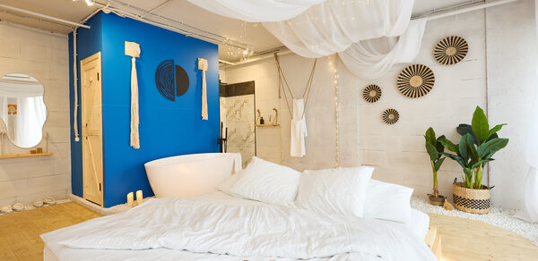 Пляжный лофт с галькой, открытой ванной, панорамными окнами. Просторная комната с белыми стенами, деревянным полом и ярко-синей акцентной стеной. Большая кровать с белым постельным бельём, рядом белая ванна. Растения в углу, потолок украшен тканью и светодиодными огнями. Декор: круглые элементы на стене.