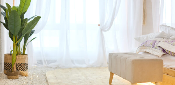 Пляжный лофт с галькой, открытой ванной, панорамными окнами. Светлое пространство с деревянным полом и белыми прозрачными занавесками. Слева стоят два плетёных горшка с зелёными растениями. Справа бежевый диван-бенч на деревянных ножках. На заднем плане видны постельные принадлежности.