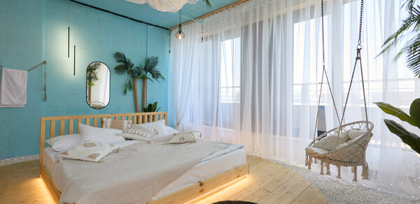 Лофт Мальдивы с открытым душем, балконом и видом на Сити. Спальня с деревянной кроватью на круглой платформе и подвесным креслом. Голубые стены, белые шторы, зеркало, небольшие растения. Натяжной светлый потолок с текстилем, светильники. Нейтральное постельное белье, коврик, белый и песочный цвета.