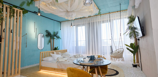 Лофт Мальдивы с открытым душем, балконом и видом на Сити. Современное праздничное пространство с голубыми стенами. Деревянная кровать с подсветкой, жёлтые кресла, круглый стол. Белые шторы на больших окнах, качели, телевизор. Потолок декорирован тканью и зеленью. Напольное покрытие из светлого дерева.