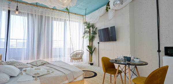 Лофт Мальдивы с открытым душем, балконом и видом на Сити. Спальня с двуспальной кроватью, столиком и двумя стульями. Голубой потолок с декоративной тканью, светлые шторы, подвесные лампы, телевизор на стене, растения в горшках. Светлое деревянное покрытие.