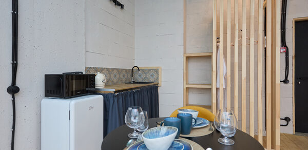 Лофт Мальдивы с открытым душем, балконом и видом на Сити. Небольшая кухонная зона с белым холодильником, микроволновой печью и чайником. Слева бетонная стена, потолок голубого цвета. Круглый стол накрыт на двоих с чашками и стаканами. Справа деревянная перегородка, рядом полка. Пол светлый.