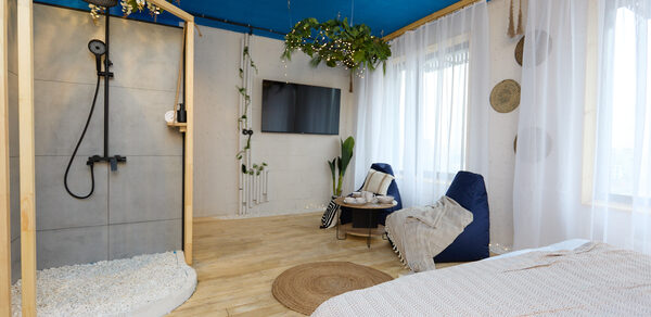 Лофт Бали с фото зоной на 23 этаже с видом на Москва-реку. Просторная комната с деревянным полом и синим потолком. Душевая зона с серыми плитками и черной сантехникой, рядом кровать с покрывалом, два кресла-мешка, телевизор на стене, прозрачные шторы. Декор из подвесных растений, коврик.