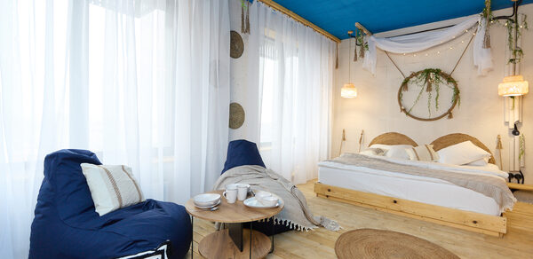 Лофт Бали с фото зоной на 23 этаже с видом на Москва-реку. Спальня с деревянным полом и синим потолком, кровать с белым постельным бельём, два кресла-мешка, круглый стол, подвесные светильники, декоративные растения и круглый ковёр. Белые занавески на окне, бежевые и черные подушки.