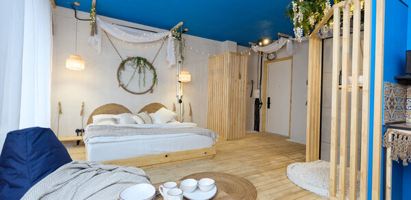 Лофт Бали с фото зоной на 23 этаже с видом на Москва-реку. Спальня с двуспальной кроватью, деревянными панелями и голубым потолком. Декор из растений, светильники, круглый стол с посудой, кресло-мешок. Светлые деревянные полы, стены декорированы тканями и венком. Гардероб с открытыми полками.