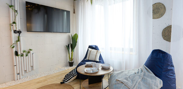 Лофт Бали с фото зоной на 23 этаже с видом на Москва-реку. Современное пространство с деревянным полом, двумя синими креслами-мешками, круглым кофейным столиком с белыми тарелками и чашками. Белая штора на окне, телевизор на стене, декоративные элементы из растений и труб, мягкое освещение.