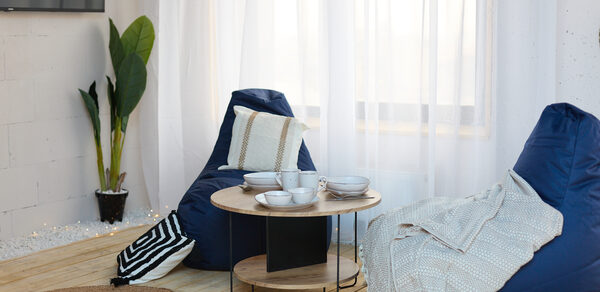 Лофт Бали с фото зоной на 23 этаже с видом на Москва-реку. Современная гостиная с двумя синими креслами-мешками, круглым деревянным столом с посудой, растением в горшке, светлыми шторами, круглым ковриком и деревянным полом. Белая кирпичная стена с телевизором, декор в нейтральных тонах.