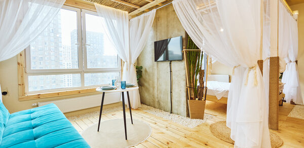 Тропический лофт с бунгало, баром и французским балконом. Жилая комната с деревянным полом и потолком, оформленным из бамбука. Синий диван, круглый столик с двумя стаканами. Светлые шторы разделяют зону с кроватью. Декор растениями в горшках. Мягкое дневное освещение из окна, телевизор на стене.