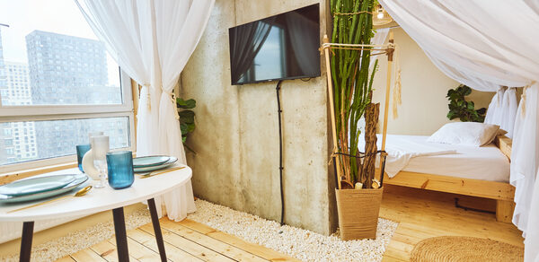 Тропический лофт с бунгало, баром и французским балконом. Светлое помещение с деревянным полом и тканевым навесом. В центре бетонная стена с телевизором. Слева стол с посудой, справа кровать. У стены большие окна, декорированы растения. Под ногами ковровая дорожка и белая галька.