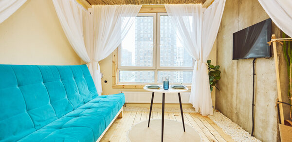 Тропический лофт с бунгало, баром и французским балконом. Просторная комната с деревянным полом и потолком. Голубой диван у стены, напротив — круглый стол с черными ножками. Белые занавески на окнах и вокруг дивана. На стене телевизор, рядом растение. Светлая, минималистичная обстановка.