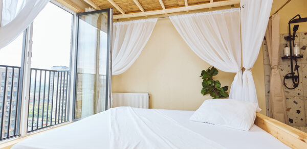 Тропический лофт с бунгало, баром и французским балконом. Спальня с двуспальной кроватью, белым постельным бельем и балдахином. Большое окно и стеклянная дверь ведут на балкон с черными перилами. Натуральный потолок с деревянными балками. На стене радиатор. В углу зелёное растение. Светлые занавеси.