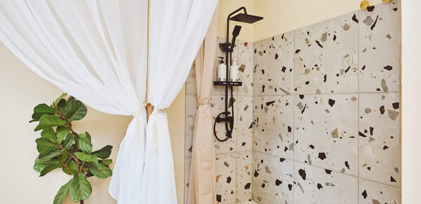 Тропический лофт с бунгало, баром и французским балконом. Душевая зона с терраццо плиткой на стене, черный душ, светлые ткани, бежевые стены, растение в горшке, деревянные элементы, стеклянные бутылочки на полке, белый текстиль.