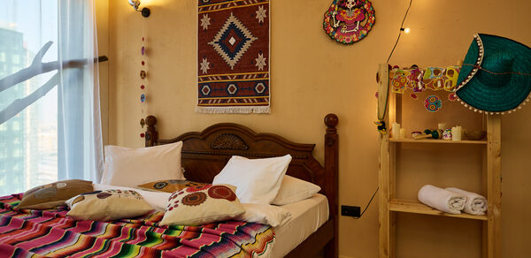⁠Лофт в Мексиканском стиле для костюмированных вечеринок. Спальня с деревянной кроватью, украшенная цветным покрывалом и подушками. Стены в бежевых тонах с декоративными элементами, полка с декоративными предметами и полотенцами. Освещение от настенных бра. Белые занавески на окне.