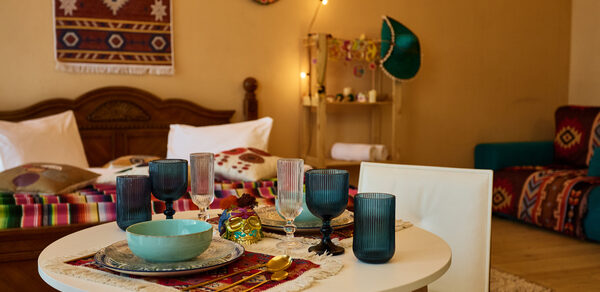 ⁠Лофт в Мексиканском стиле для костюмированных вечеринок. Гостиная с деревянным столом, белыми стульями и сервировкой из цветных бокалов и тарелок. На стене текстильный декор, рядом кровать с разноцветным покрывалом, в углу диван с ярким пледом. Теплое освещение и деревянный пол.